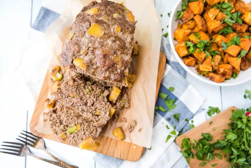 Sweet-and-sour AIP meatloaf sliced and resting on a wooden cutting board - another horizontal view from directly above with a bowl of roasted diced sweet potatoes on the side and fresh parsley in the corner