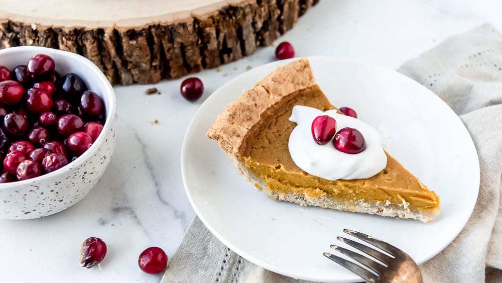 A side view of a slice of pumpkin pie with a topping of whipped cream and fresh cranberries