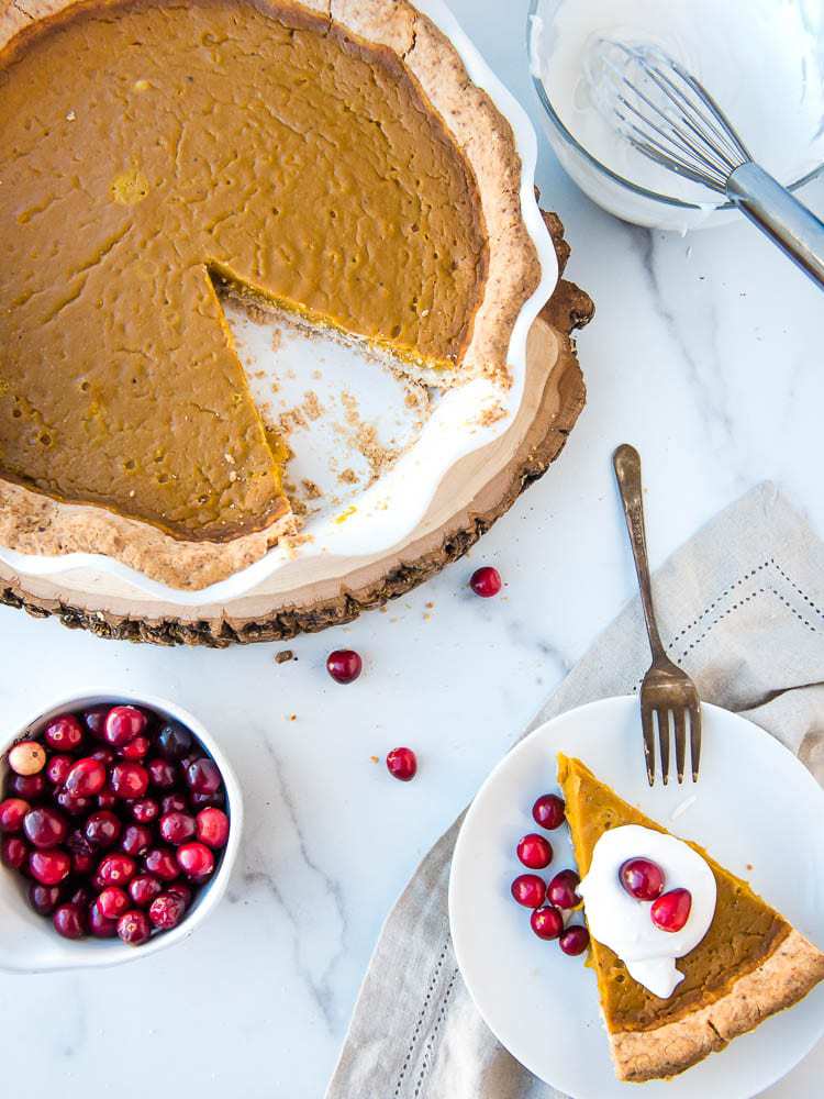 An overhead view of a slice of pumpkin pie with a topping of whipped cream and fresh cranberries and the rest of the pumpkin pie as well