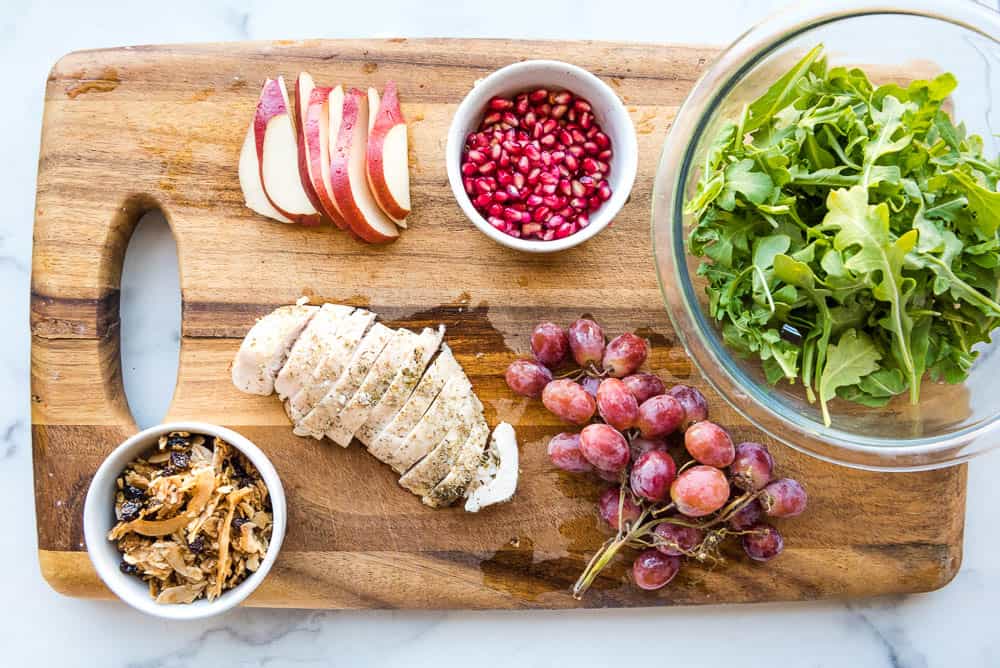 Overhead view of ingredients on a cutting board, from top left going clockwise: sliced pears, pomegranate arils, arugula in glass mixing bowl, red graps, baked chicken breast, white cup filled with cinnamon raisin tigernut granola