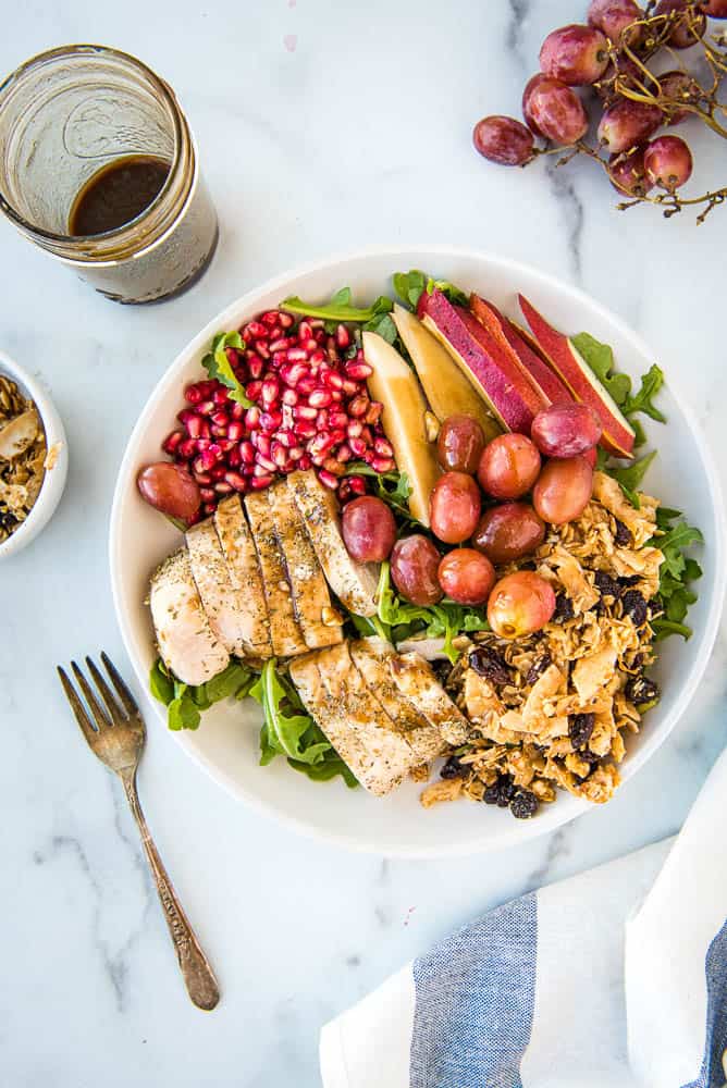 Overhead view of completed pear salad with pomogranete arils, red pears, grapes, cinnamon raisin tigernut granola, sliced baked chicken breast on a bed of arugula in a white bowel.