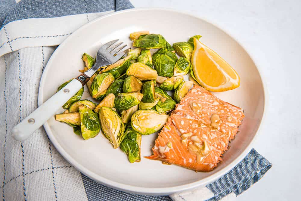 Closeup view of a piece of salmon, Brussles sprouts and a lemon wedge on a white plate with a fork on top ofa blue and white checkered napkin
