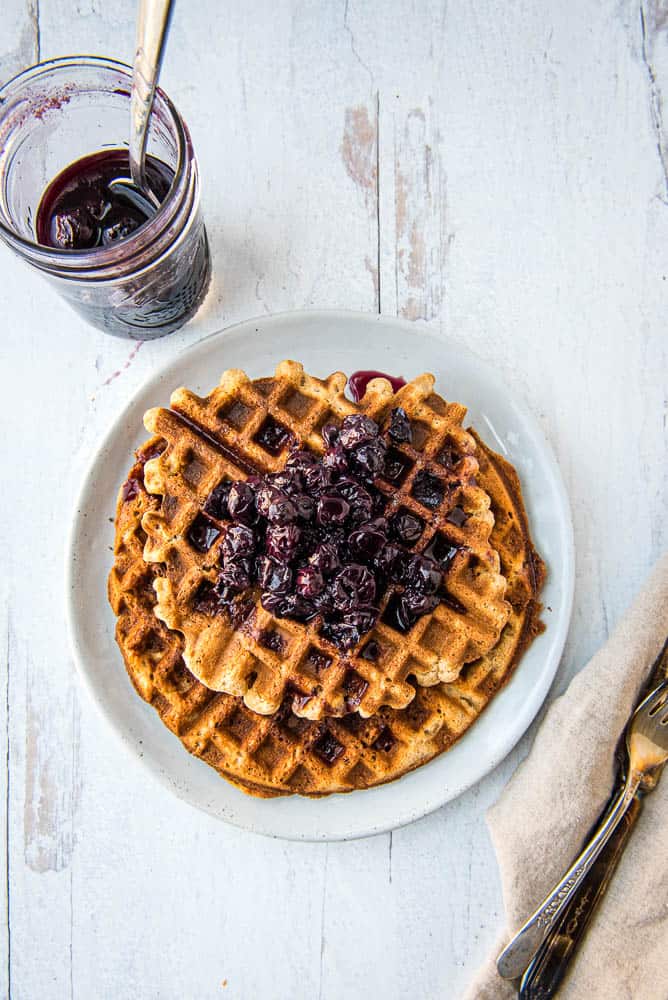 Overhead view of two tigernut waffles with blueberry syrup stacked on white plate with a jar or blueberry syrup and a silver spoon in upper left quadrant and utinsels on a napkin in lower right quadrant