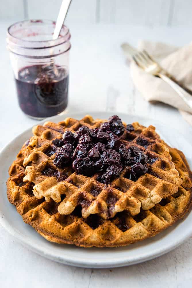 Eye level angle of two tigernut waffles stacked on a white plate with a jar of blueberry syrup and a fork on a napkin in the background.