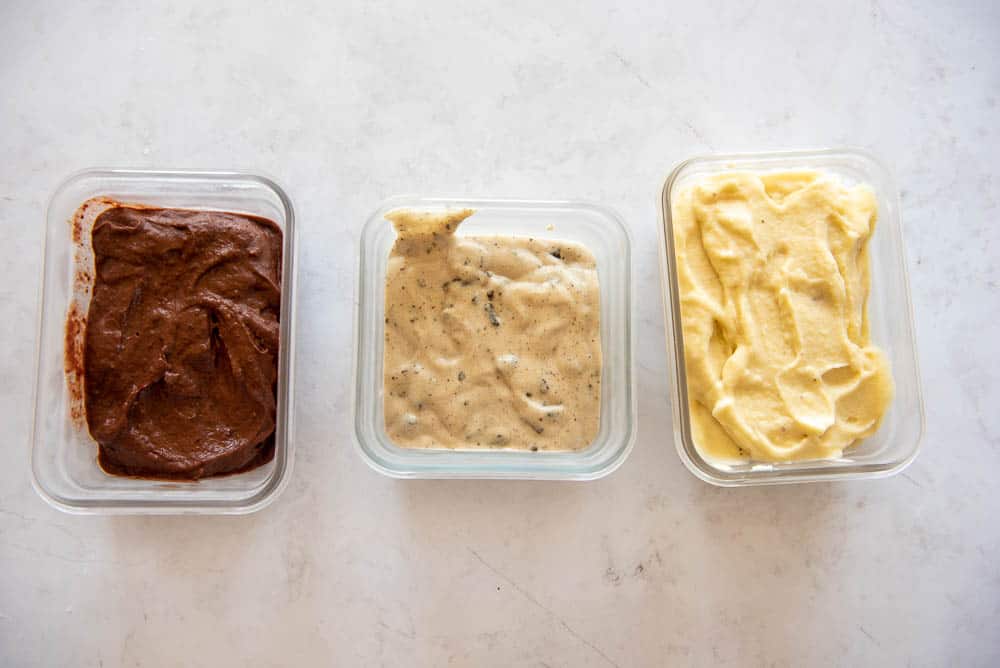 An overhead view of three square shaped glass containers, filled with three different AIP Ice Cream flavors; on the left is a container of Pina Colada Ice Cream, in the middle is a container with Chocolate Chip Ice Cream and on the left a container of Cherry Chocolate Ice Cream.