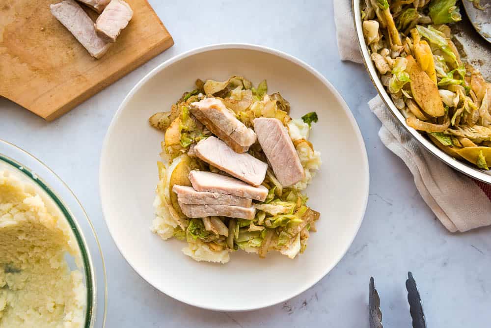Overhead view of a white plate in the center of the table with mashed cauliflower, sautéed apples, and slices of pork stacked on top of each other in that order. In the upper left corner, there is a cutting board with slices of pork. In the top right corner, there is a pan resting onto pf a beige towel with sautéed cabbage inside the pan. In the bottom left, there is a glass bowl filled with mashed cauliflower. In the bottom right-center, there are two stainless steel pincers from some tongs.