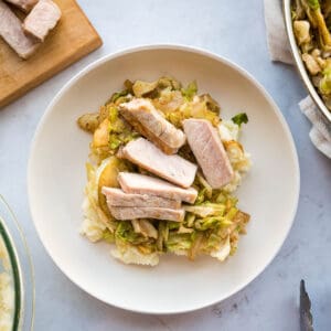 Overhead view of a white plate in the center of the table with mashed cauliflower, sautéed apples, and slices of pork stacked on top of each other in that order. In the upper left corner, there is a cutting board with slices of pork. In the top right corner, there is a pan resting onto pf a beige towel with sautéed cabbage inside the pan. In the bottom left, there is a glass bowl filled with mashed cauliflower. In the bottom right-center, there is one stainless steel pincer from some tongs.