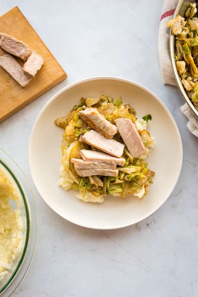Overhead view of a white plate in the center of the table with mashed cauliflower, sautéed apples, and slices of pork stacked on top of each other in that order. In the upper left corner, there is a cutting board with slices of pork. In the top right corner, there is a pan resting onto pf a beige towel with sautéed cabbage inside the pan. In the bottom left, there is a glass bowl filled with mashed cauliflower.