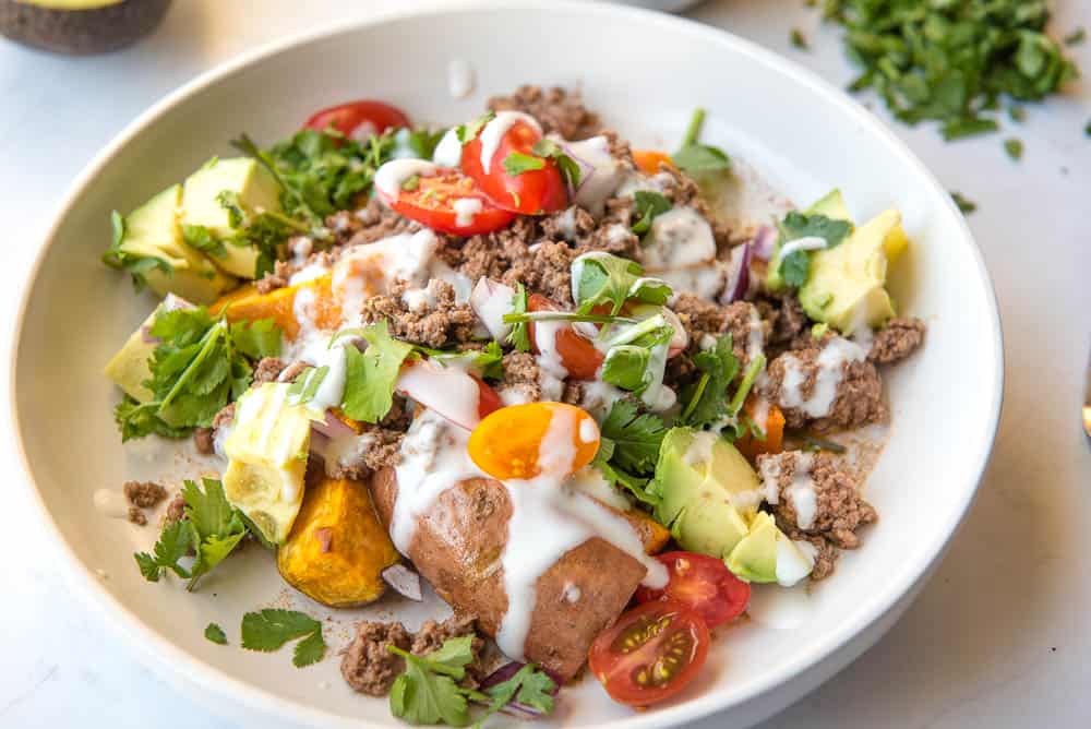 Side view of white bowl filled with roasted chopped sweet potatoes, halved cherry tomatoes, chopped avocado, cooked ground beef, chopped cilantro, with sour cream drizzled on top.