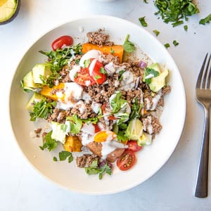 Overhead view of white bowl filled with roasted chopped sweet potatoes, halved cherry tomatoes, chopped avocado, cooked ground beef, chopped cilantro, with sour cream drizzled on top. The bowl is next to a stainless steel fork to the right, half of an avocado in upper left quadrant, and chopped cilantro to the top right quadrant.