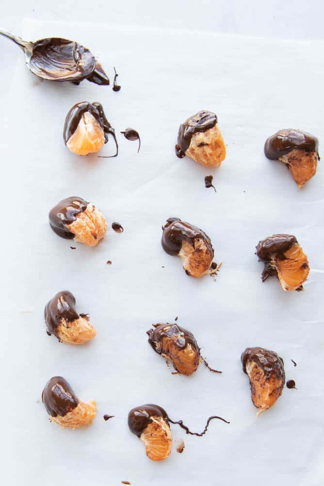 Overhead view of a piece of white parchment paper with a chocolate covered spoon in the upper left hand corner and a grid of 3 across and 4 down of orange slices dipped in chocolate.